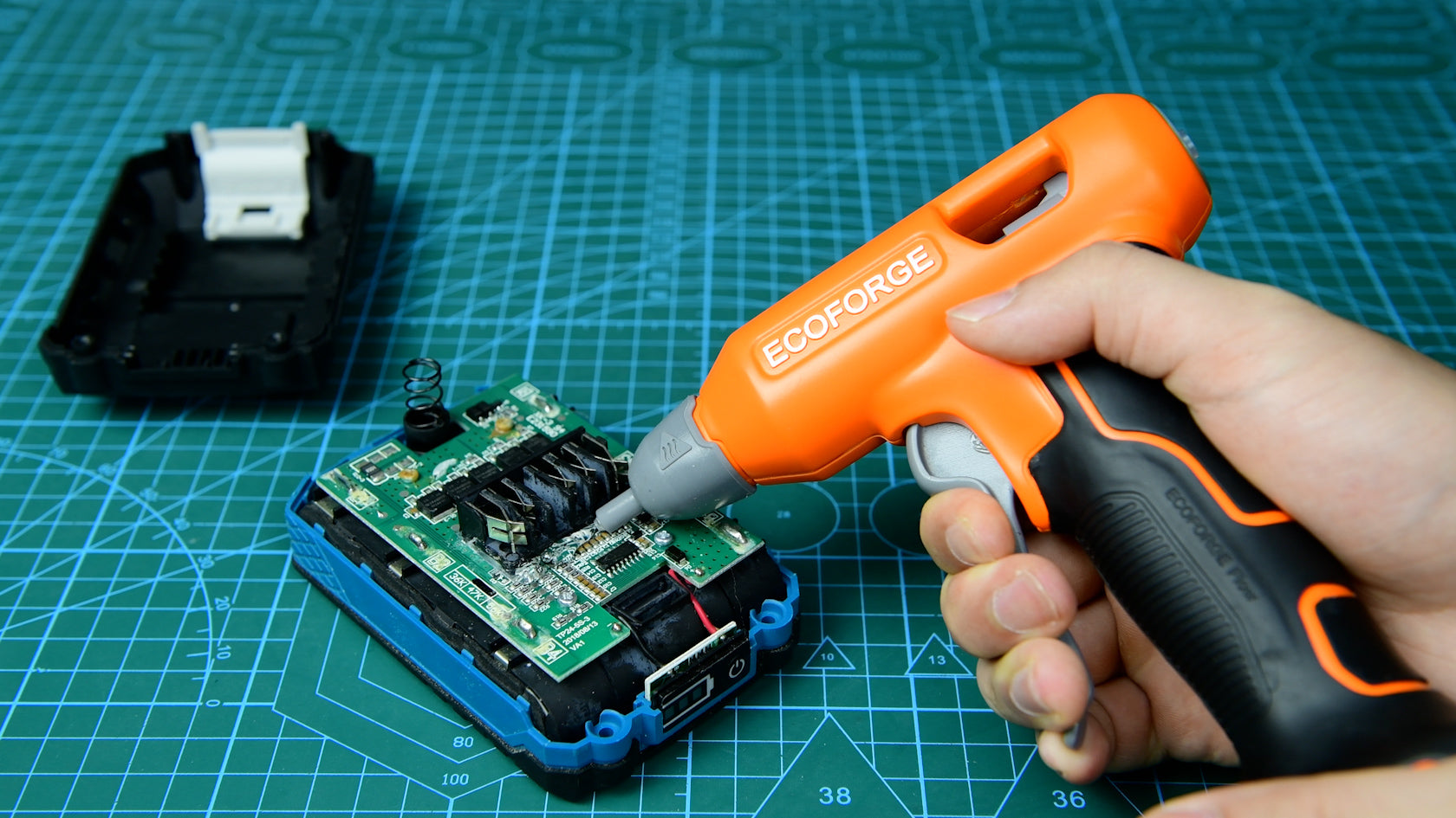 A close-up photograph of a person using an orange and black ECOFORGE cordless hot glue gun to apply adhesive to components on an exposed circuit board, set on a green grid cutting mat.