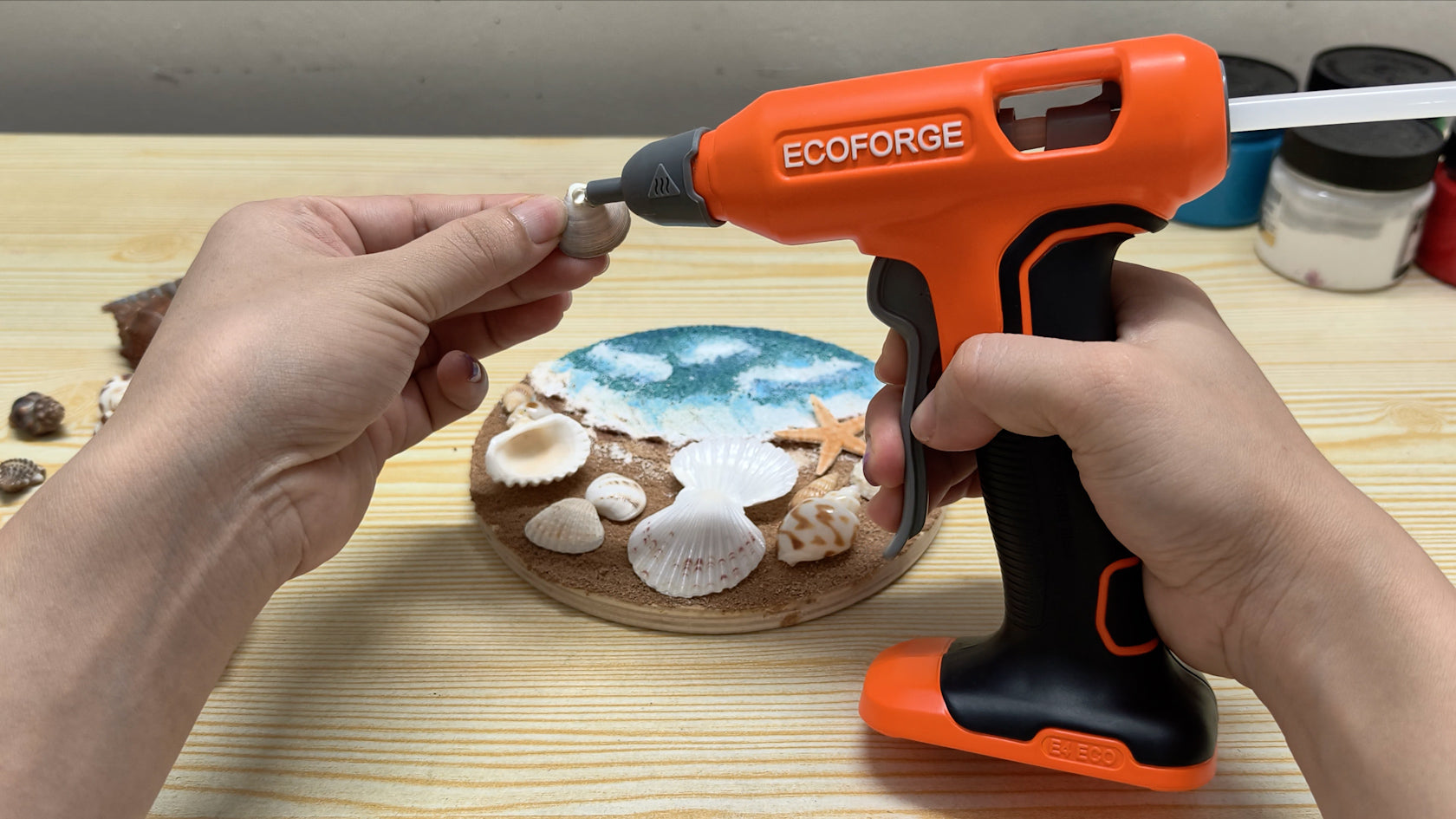 A close-up photograph of a person using an orange and black ECOFORGE cordless hot glue gun to apply adhesive to a small seashell, crafting a beach-themed round wooden coaster on a light wood table.