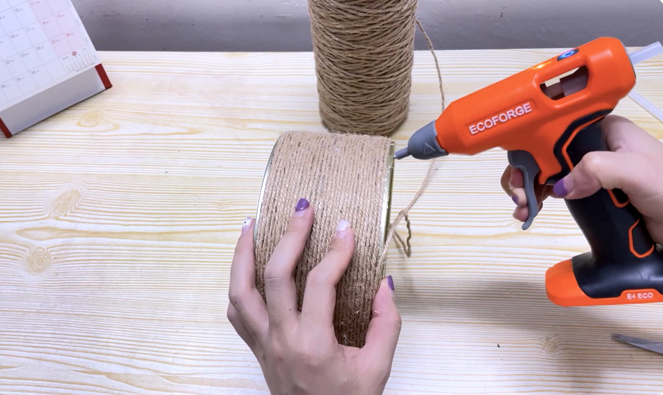 A close-up photograph of a person using an orange and black ECOFORGE cordless hot glue gun to secure natural jute twine around a metal tin, set on a light wood workbench with a spool of rope in the background.