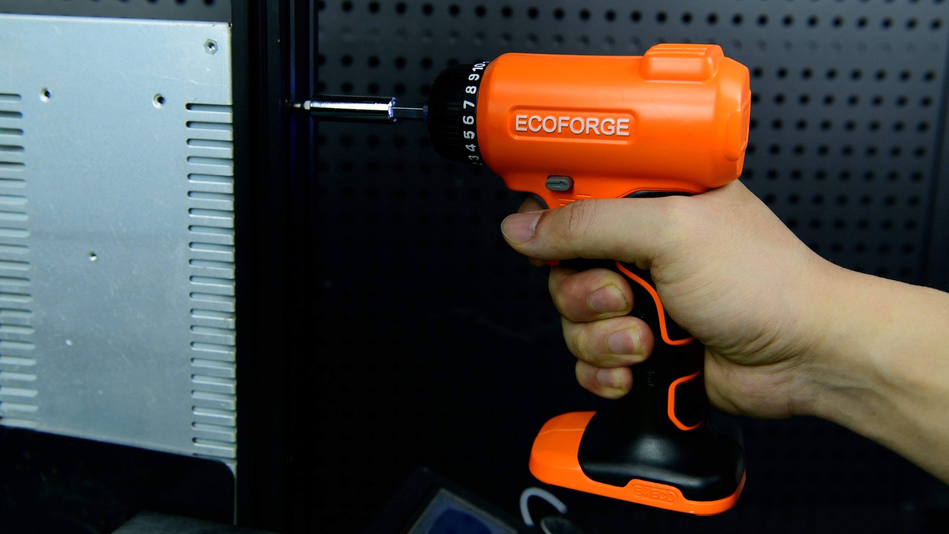 A photograph of a person holding an orange and black ECOFORGE electric screwdriver, using a bit extension to drive a screw into a metal equipment frame against a dark pegboard background.