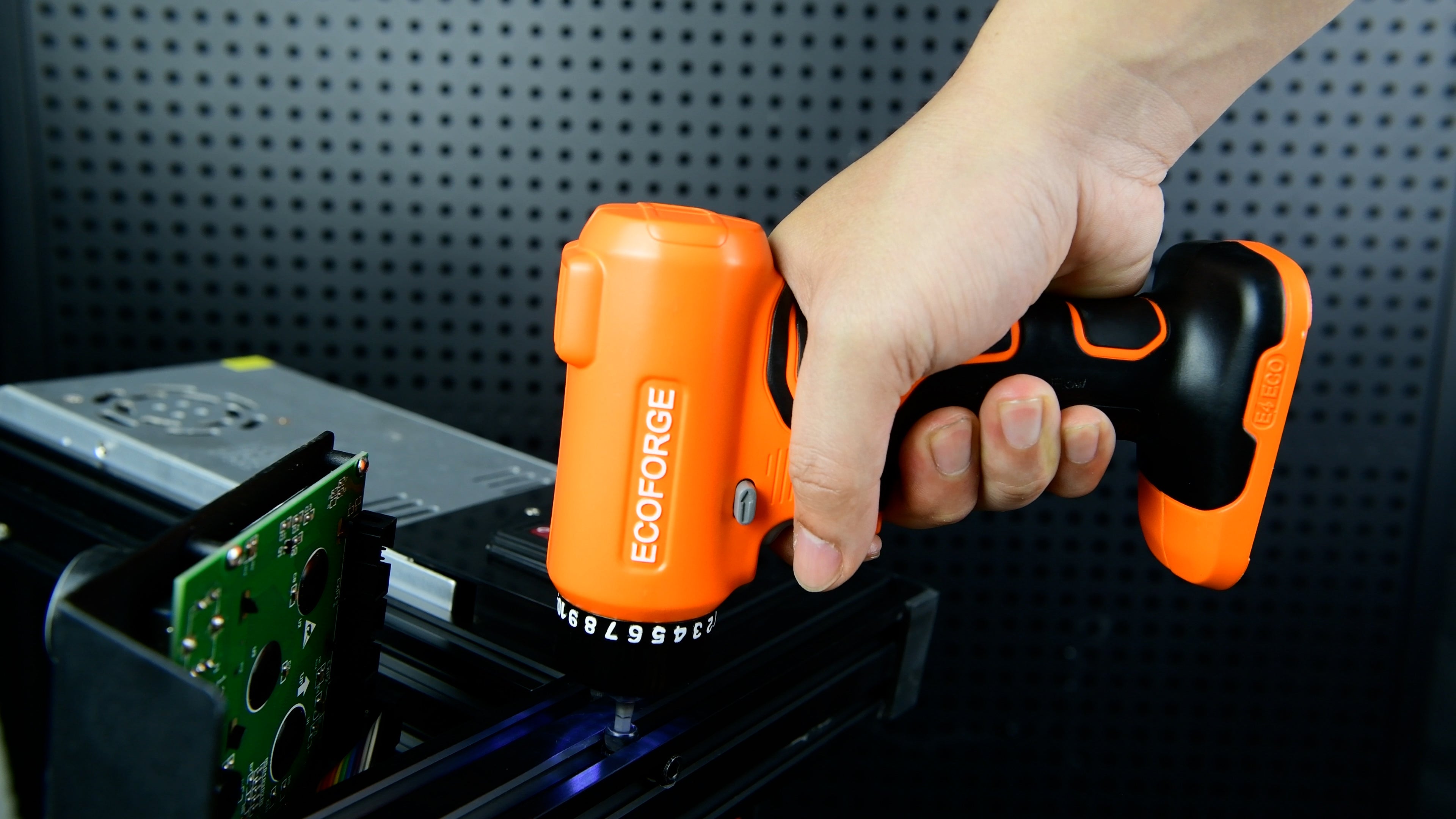 A photograph of a person using an orange and black ECOFORGE electric screwdriver to drive a screw vertically into a black aluminum extrusion rail near electronic components, set against a dark pegboard background.