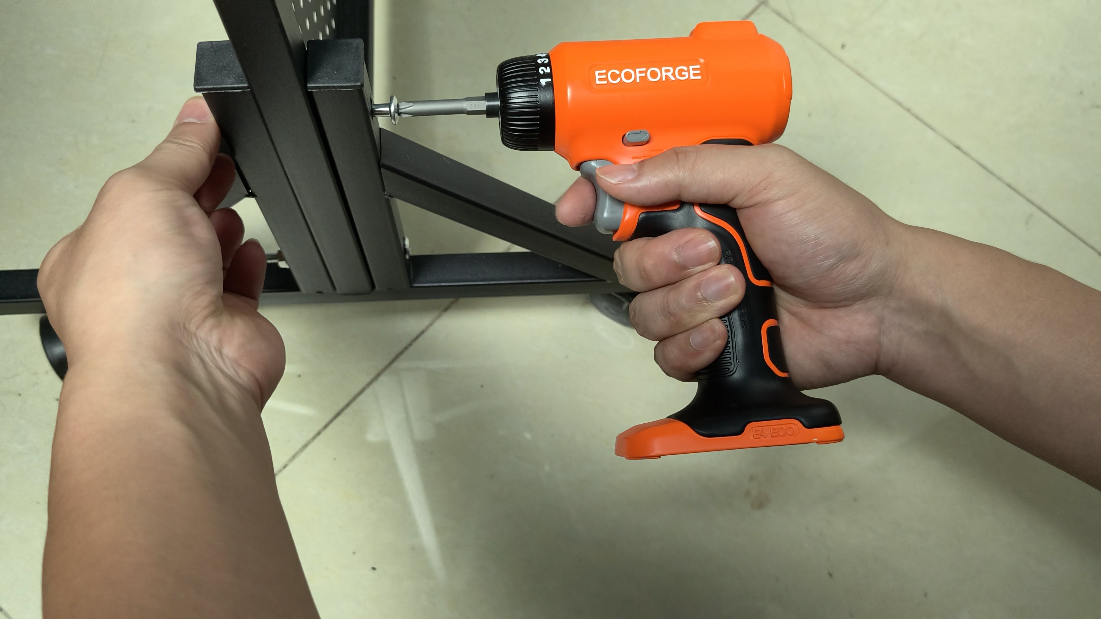 A photograph of a person using an orange and black ECOFORGE electric screwdriver to drive a screw into a black metal frame during furniture assembly, set against a light-colored tiled floor.