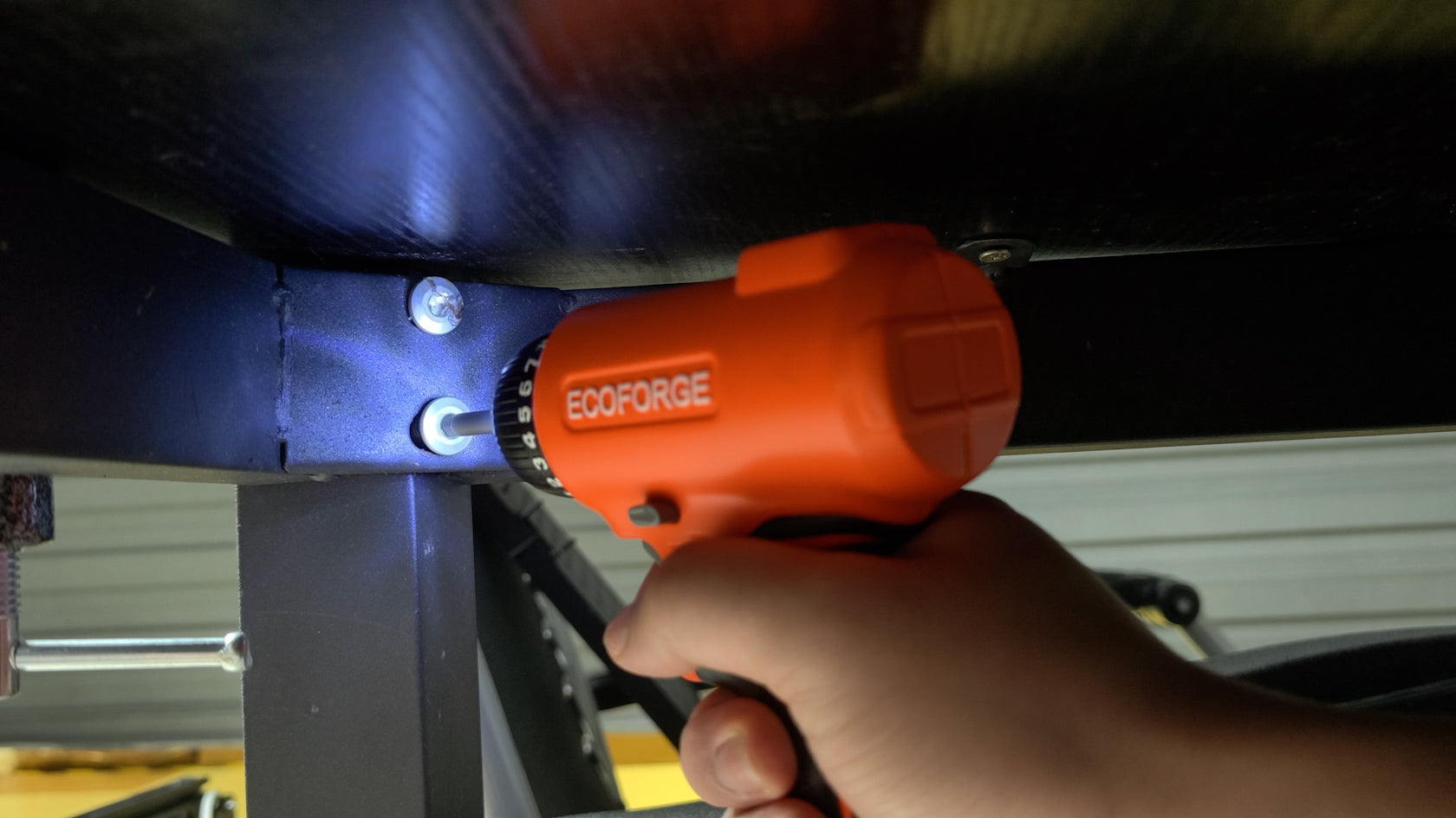 A photograph of a person holding an orange ECOFORGE electric screwdriver, utilizing its built-in LED light to illuminate and drive a screw into a black metal frame joint in a dim workspace under a desk.
