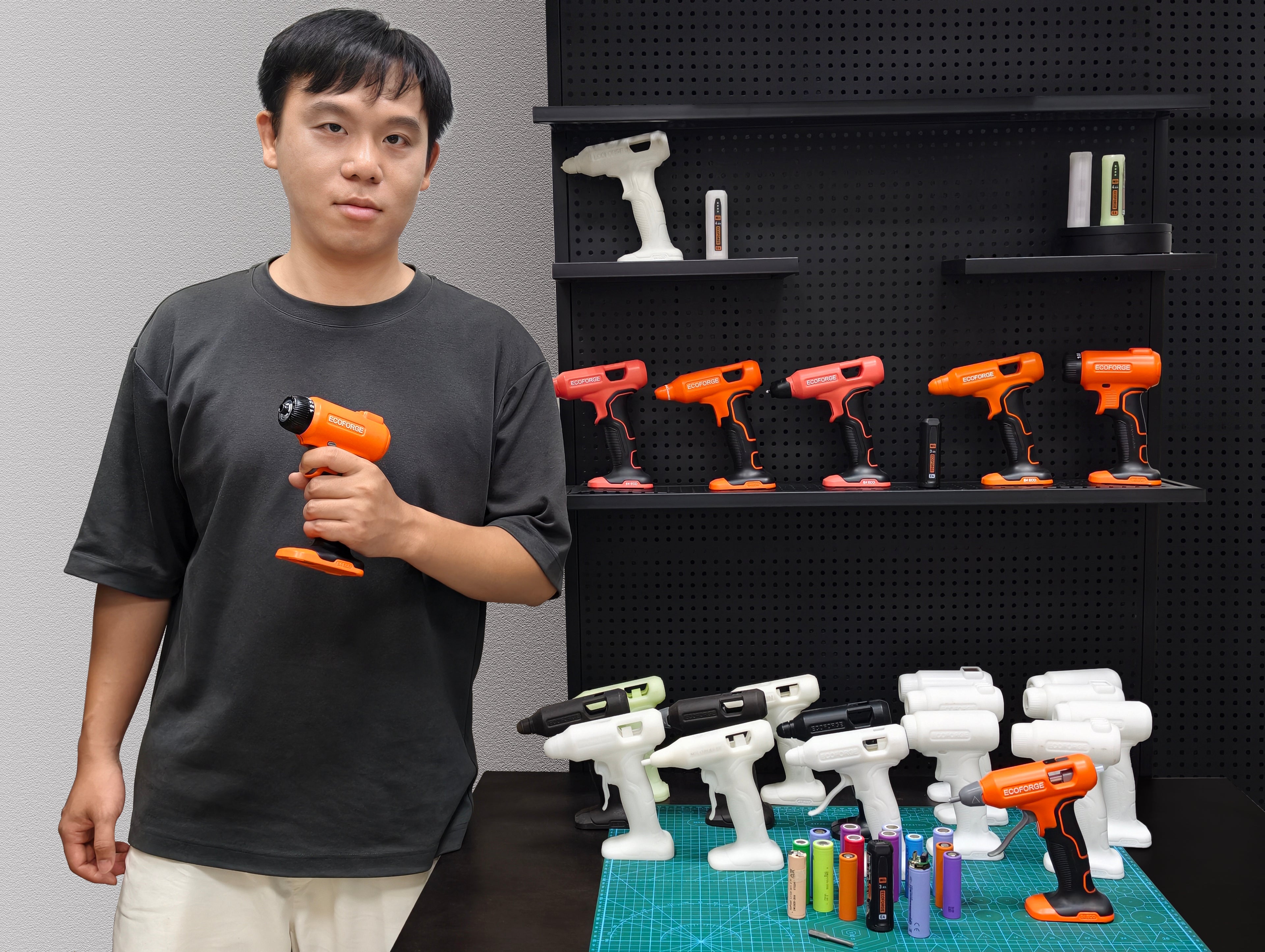 A portrait photograph of a person holding an orange ECOFORGE electric screwdriver, standing in a workshop beside a black pegboard shelf displaying tool design iterations and a workbench arranged with white 3D-printed prototypes and various lithium-ion battery cells.