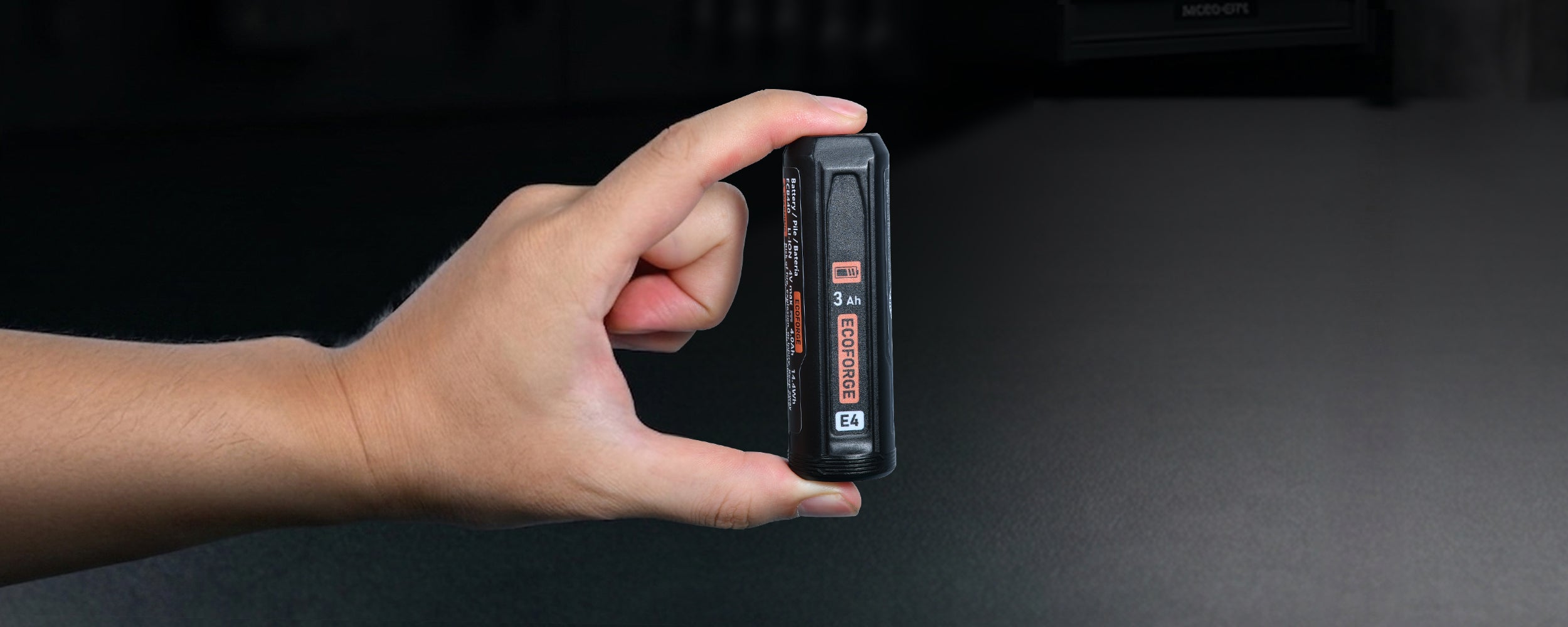 A close-up photograph of a hand holding a compact, cylindrical black ECOFORGE E4 battery pack vertically between a thumb and index finger. The battery features an orange battery icon, "3 Ah" capacity labeling, a vertical orange ECOFORGE logo, and a white "E4" badge near the base. The background shows a dark, softly blurred workshop setting with tools mounted on a wall board, emphasizing the battery's lightweight and portable size.
