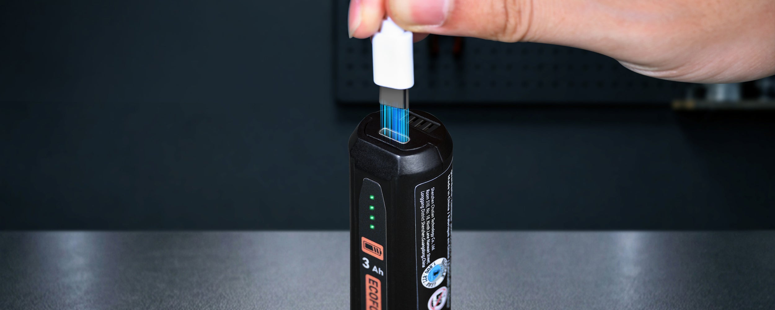 A close-up photograph of a hand plugging a white USB-C cable into the top port of a black ECOFORGE E4 battery pack. Stylized glowing blue light lines extend from the connector into the port to represent charging power flow. On the side panel of the battery, four green LED indicator lights are illuminated above an orange battery symbol and "3 Ah" capacity label, against a dark, blurred background.