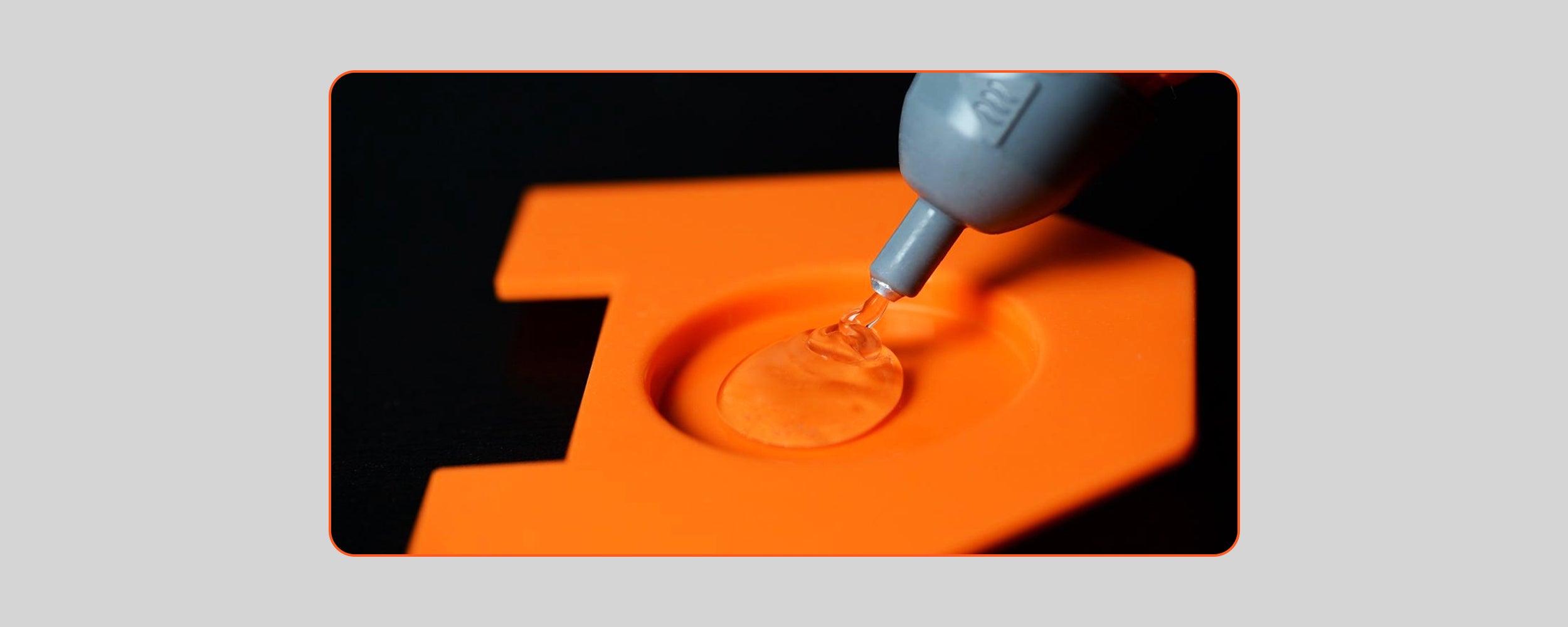 An extreme close-up shot of the extended grey precision nozzle of a hot melt glue gun dispensing a bead of clear, melted glue into a small circular recessed area on a bright orange plastic workpiece