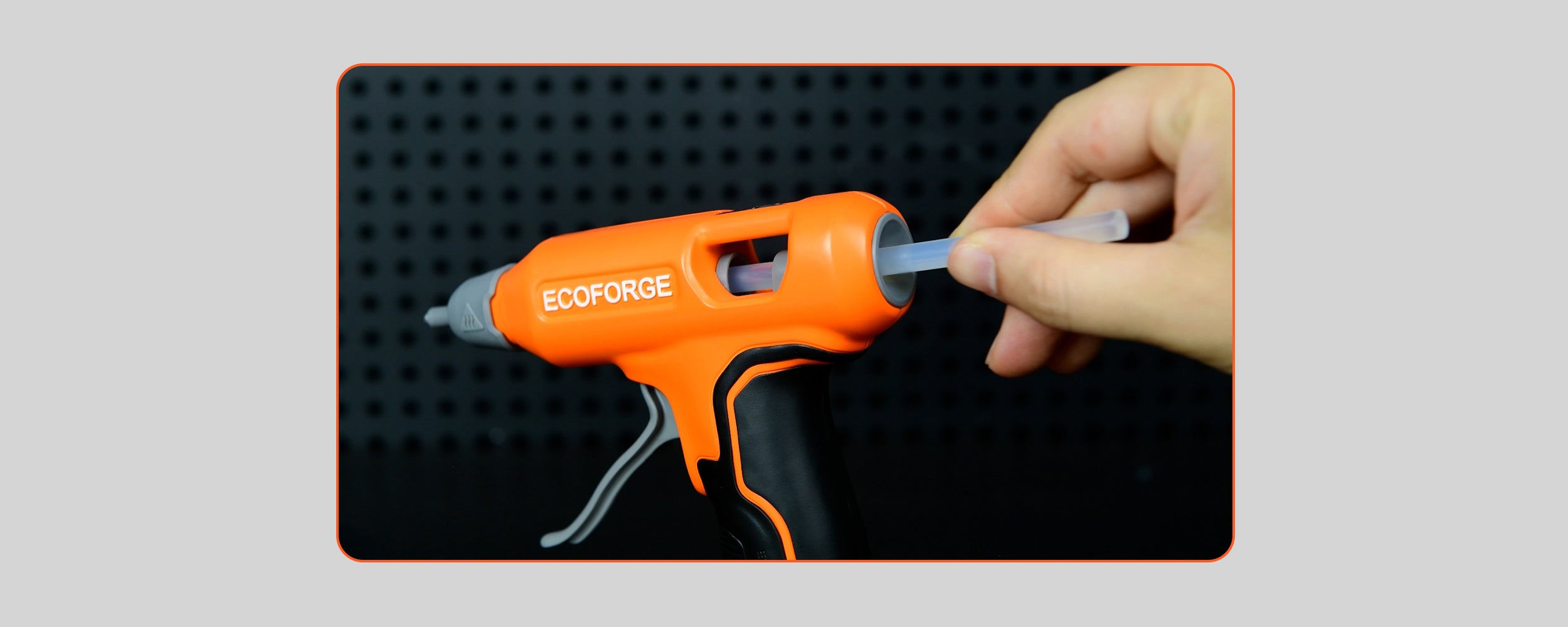 A close-up photograph of a hand inserting a clear glue stick into the rear entry port of the orange ECOFORGE hot glue gun. The inlet port features a dark grey funnel-shaped ring designed for smooth loading, set against a dark pegboard background.