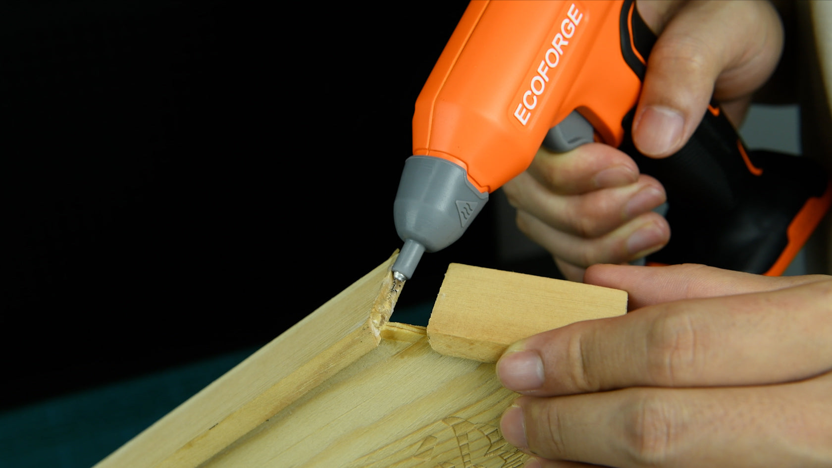 A close-up photograph of a person using an orange and black ECOFORGE cordless hot glue gun to apply adhesive to a wooden joint. One hand grips the ergonomic tool while the other holds a small wooden piece in place against a dark background.